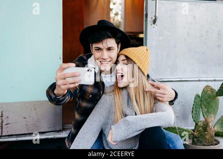 Hübscher junger Mann und schöne Frau, die lächeln und für Selfie posieren, während sie in der Nähe eines verwitterten Wohnwagens sitzen Stockfoto