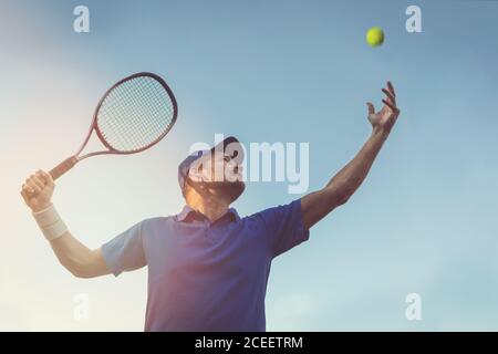 Aktiver junger Mann spielt Tennis im Freien. Servieren Sie den Ball gegen blauen Himmel Stockfoto