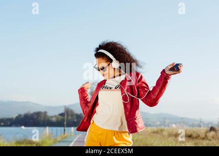 Schöne afroamerikanische Frau in roter Jacke, die gute Musik hört und tanzt, während sie an sonnigen Tagen entlang der Landstraße spazierengeht Stockfoto