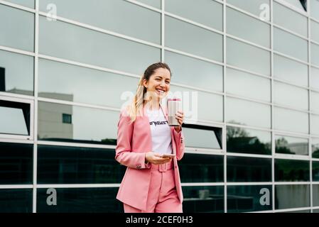 Junge stilvolle Frau hält Tasse mit Kaffee und Smartphone lachend vor der Kamera auf der Straße. Stockfoto
