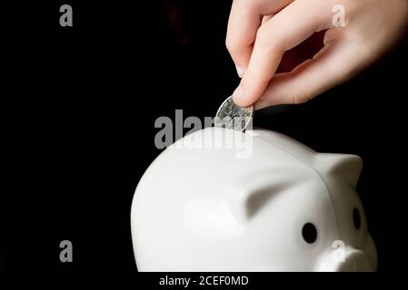 Hand des Kindes, das ein Viertel in ein Sparschwein legt. Sparkonzept, schwarzer Hintergrund Stockfoto