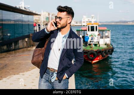 Stilvoller schöner Mann in Denim und Jacke mit Telefonanruf, während er mit Lederrucksack am Meer steht. Stockfoto