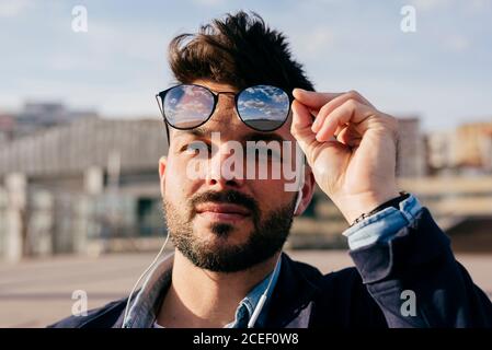 Kopfaufnahme eines trendigen bärtigen Mannes mit Frisur über Kopfhörer und Sonnenbrillen, die im Sonnenlicht wegschauen. Stockfoto