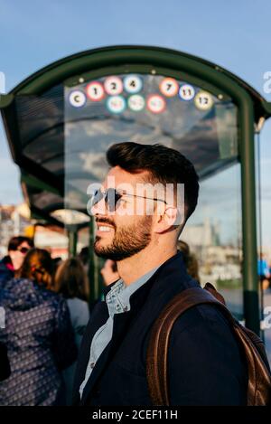 Seitenansicht des trendigen bärtigen Mannes in Sonnenbrille, der mit Rucksack im Sonnenlicht auf der Reisestation steht und lächelt. Stockfoto