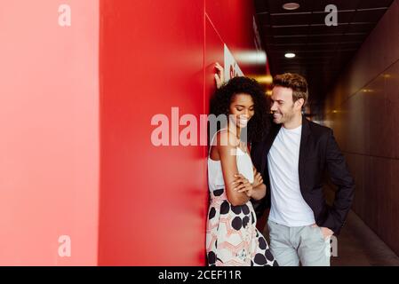 Hübscher junger Kerl, der sich an der roten Wand lehnt und mit ihm flirtet Schöne afroamerikanische Frau, während sie im Gebäude Korridor zusammen stehen Stockfoto