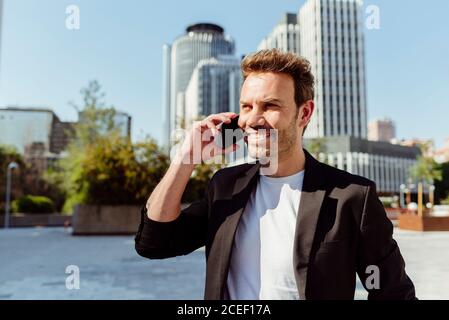 Attraktiver junger Mann lächelt mit dem Smartphone, während er auf der Straße steht Der modernen Stadt an sonnigen Tag Stockfoto