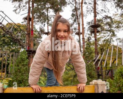 Glücklich lächelndes Teenager-Mädchen in einem Kunstpelzmantel und mit langem Pferdeschwanz klettern den gelben Kletterer auf dem Kinderspielplatz. Seilpark mit Pinien Stockfoto