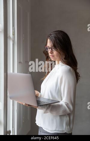 Attraktive Erwachsene Frau in elegantem Outfit in der Nähe von Büro-Fenster stehen Und das Surfen in einem modernen Laptop Stockfoto