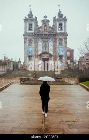 Rückansicht des Weiblichen mit weißer Schirm wandern auf alten Pflaster auf die schöne Kirche Gebäude an regnerischen Tag in Porto, Portugal Stockfoto