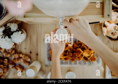 Oben Schuss der Hände der anonymen Frau, die schön ausgießt Getränk aus dem Wasserhahn über Holztisch während der Party im Garten Stockfoto