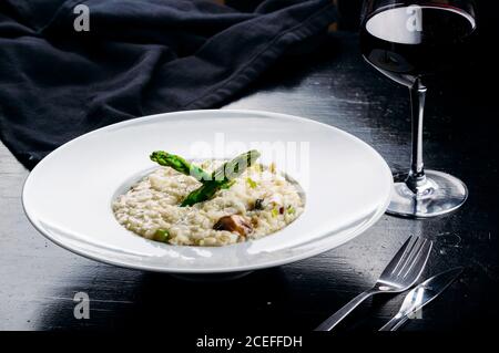 Nahaufnahme eines wunderschön servierten weißen Porzellantellers mit Risotto und Gemüse auf einem dunklen Tisch mit Gabel, Messer und Glas Wein Stockfoto