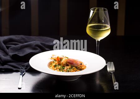 Elegante weiße Keramikplatte mit hellem, köstlichem Risotto mit Garnelen Platziert auf dunklem Holztisch mit Gabel und Glas Weißwein Stockfoto