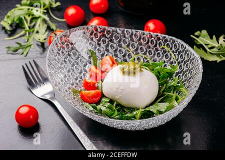 Nahaufnahme einer schönen Glasschüssel mit gehackten Kirschtomaten, Potherbs und köstlicher Ziegenkäse-Kugel auf dunklem Tisch mit Gabel und frischem Gemüse Stockfoto