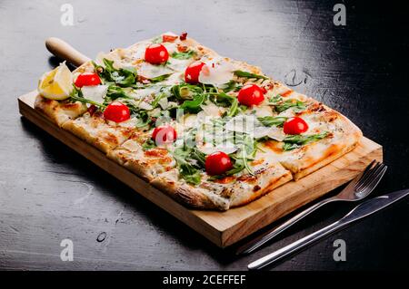 Serviert auf Holzbrett frische rechteckige geschnitten Pizza mit Kirschtomaten, Potherbs und Käse auf dunklen Tisch mit verstreuten Mehl, Gemüse, Gabel und Pizzaschneider von oben platziert Stockfoto