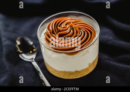 Nahaufnahme von cremigem Dessert in Tasse komponiert und mit köstlichen Sahne serviert auf schwarz mit Löffel gekrönt. Stockfoto
