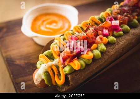 Nahaufnahme des servierten Hot Dogs mit schwarzem Brot Brötchen mit verschiedenen Saucen und Zusatzstoffen auf dem Teller serviert gekrönt. Stockfoto