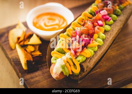 Nahaufnahme des servierten Hot Dogs mit schwarzem Brot Brötchen mit verschiedenen Saucen und Zusatzstoffen auf dem Teller serviert gekrönt. Stockfoto