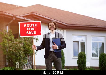 Joyful Immobilienmakler Holding Zeichen HAUS ZUM VERKAUF in der Nähe Wohngebäude im Freien Stockfoto