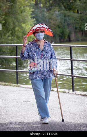 Eine stilvolle Frau in ihren Siebzigern geht für einen Übungsspaziergang mit einer chirurgischen Maske & passendem Hut & Geldbörse. In einem Park in Flushing, Queens, New York. Stockfoto