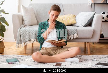 Konzentrierter Teenager Junge sitzt auf dem Boden im Zimmer, Buch zu lesen Stockfoto