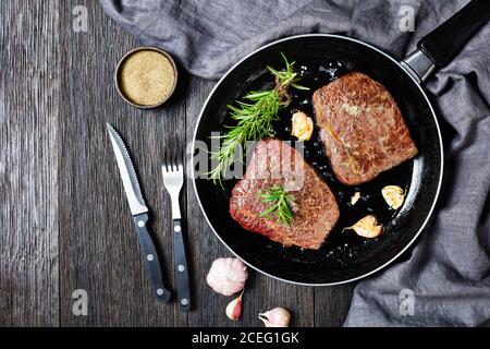 Gebratene Rindersteaks mit Rosmarinstrauß, Knoblauch und Salz auf einer Pfanne auf einem Holztisch mit Gabel und Messer, horizontale Ansicht von oben, flach liegend, fre Stockfoto