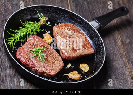 Gebratenes Rumpsteak mit Rosmarinstrauß, Knoblauch und Salz auf einer Pfanne auf einem dunklen Holztisch, horizontale Ansicht von oben Stockfoto