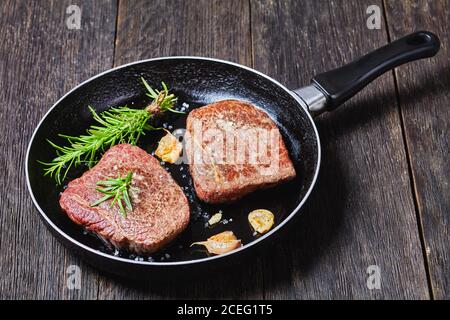 Gebratenes Rumpsteak mit Rosmarinstrauß, Knoblauch und Salz auf einer Pfanne auf einem dunklen Holztisch, horizontale Ansicht von oben Stockfoto