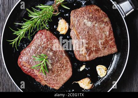 Gebratenes Rumpsteak mit Rosmarinstrauß, Knoblauch und Salz auf einer Pfanne auf einem dunklen Holztisch, horizontale Ansicht von oben, flach liegend, Nahaufnahme Stockfoto