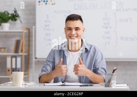 Fernlernsprache. Lächelnder junger männlicher Lehrer zeigt Daumen nach oben und erklärt den Schülern die englischen Regeln Stockfoto