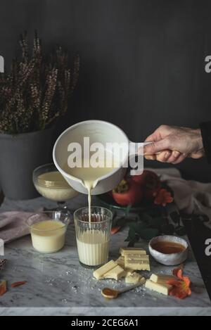 Hand der anonymen Person, die Panna cotta in Behälter in der Nähe gießt Stücke von weißer Schokolade und Honig Stockfoto