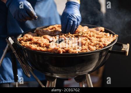 Gesichtsloser Schuss des Mannes in den Handschuhen, die rohes Fleisch anziehen Metallgitter im Grill stehen im Sonnenlicht Stockfoto
