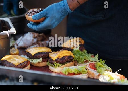 Gesichtsloser Schuss Koch in Handschuhen anordnen Brötchen mit frischen Gemüse- und Fleischpasteten mit Käseburgern Stockfoto