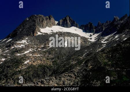 Schneefelder verweilen im Sommer inmitten der zerklüfteten Gipfel der Urner Alpen im Kanton Wallis, Schweiz, oberhalb des 7.65 km (4.75 mi) langen Rhône-Gletschers, der unterhalb des Furka-Passes auf einer Höhe von etwa 2,208 m (7,244 ft) schmilzt. Das Eis fließt mit etwa 30 bis 40 m (100 bis 130 ft) pro Jahr zu einem Schmelzwassersee, von dem aus ein Wildbach in das Talbecken stürzt. Von dort aus beginnt der Fluss Rhône seine 813 km (505 Meilen) Reise durch Südfrankreich zum Camargue Delta und dem Mittelmeer. Stockfoto