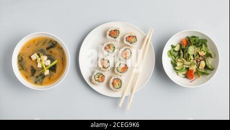 Schöne Schüsseln mit Miso-Suppe und frischem Gemüse Salat und Teller mit Sushi-Brötchen und Holzstäbchen liegen Auf weißem Hintergrund von oben Stockfoto