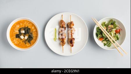 Schöne Schüsseln mit Miso-Suppe und frischem Gemüse Salat und Teller mit Sushi-Brötchen und Holzstäbchen liegen Auf weißem Hintergrund von oben Stockfoto