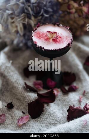 Schöne nordische Getränk in kleinen hohen Stiel Glas bestreut mit Rotes Pulver und Blatt serviert auf grauem Tuch mit gestreut Getrocknete Rosenblätter auf verschwommenem Hintergrund Stockfoto