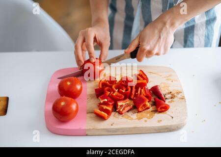 Ernte Ansicht von?Frau in Sommerkleidung Schneiden helle leckere frische Tomaten auf Holzbrett mit anderen Zutaten Kochen auf dem Tisch in der Nähe liegen Stockfoto