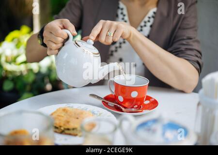 Crop-Ansicht der weiblichen Holding Porzellan Teekanne und Gießen heiß Tee in rote Keramik-Polka-gepunktet?Tasse mit Untertasse und Löffel stehen Auf weißem Tisch mit Stück Kuchen auf Teller in der Nähe Stockfoto