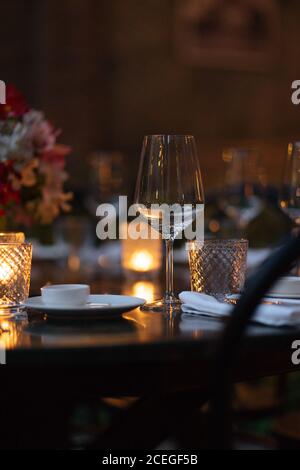 Zarte Weingläser auf dunklen Holztisch mit beleuchtet platziert Kerzen und Blumenstrauß in der Mitte am Abend mit schön Blumen und Bäume in der Umgebung Stockfoto