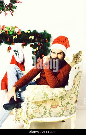Schöner bärtiger Mann mit langem Bart in weihnachten roten Hut und Pullover sitzt auf beigen Vintage-Sessel rund um weihnachtsdekoration auf weißem Studio Hintergrund Stockfoto