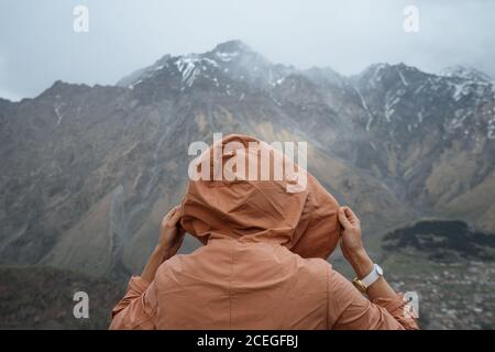 Rückansicht des Mannes in warmer Kleidung mit Rucksack Wandern In Bergen, die auf Gras stehen und auf den zerklüfteten Bergrücken blicken Bedeckt mit Schnee und Gipfeln in Wolken versteckt Stockfoto