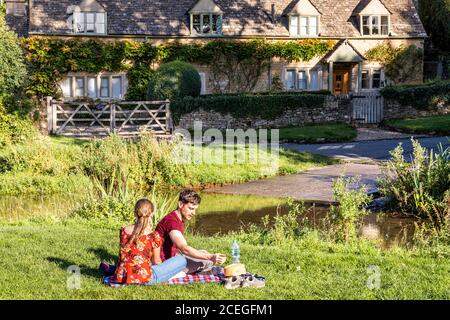 Ein junges Paar, das die Abendsonne neben der furt über dem River Eye im Cotswold Village of Upper Slaughter, Gloucestershire UK, genießt Stockfoto