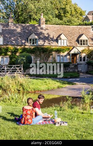 Ein junges Paar, das die Abendsonne neben der furt über dem River Eye im Cotswold Village of Upper Slaughter, Gloucestershire UK, genießt Stockfoto