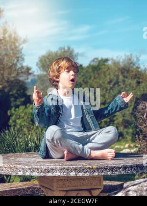 Entzückender kleiner Junge, der an sonnigen Tagen in Lotushaltung sitzt und meditiert. Stockfoto