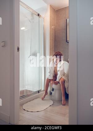 Junge im Bademantel sitzt auf der Toilette mit Zeitung Stockfoto