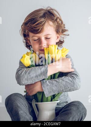 Kleiner Junge mit geschlossenen Augen sitzend und umarmt Bündel von gelben Tulpen auf grauem Hintergrund. Stockfoto