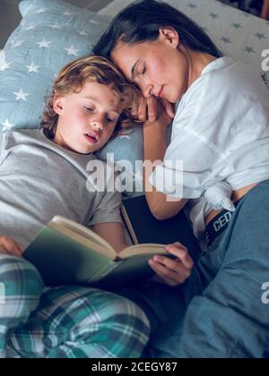 Oben ist der Schuss des süßen Jungen, der auf dem schönen Bett liegt und das interessante Buch mit Hilfe der Mutter liest. Stockfoto