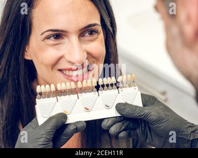 Lächelnde Frau, die einen nicht erkennbaren Zahnarzt ansieht, der im Schrank einen Prothesenfarbstoff wählt Stockfoto