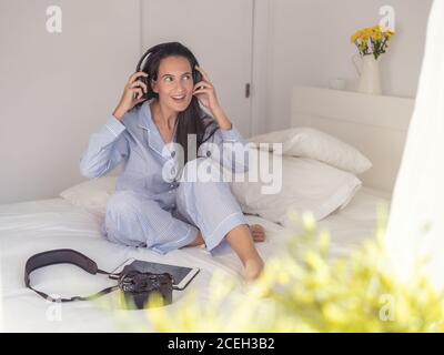 Schöne Frau trägt blauen Schlafanzug und Kopfhörer sitzen auf dem Bett, genießen Musik und wegschauen Stockfoto