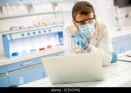 Hübscher junger Forscher in weißem Labormantel und Schutzmaske Arbeiten mit einem Laptop, während Sie im Labor sitzen Stockfoto
